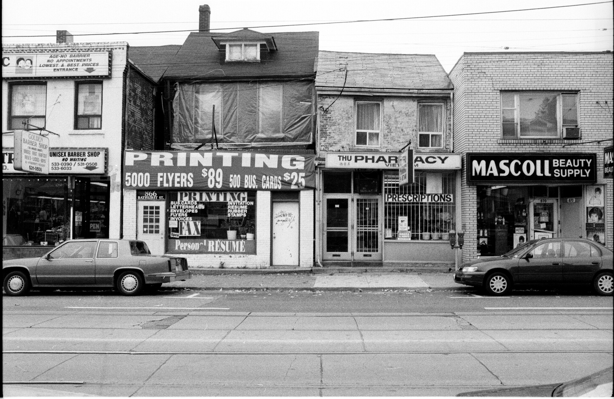 Image of four small storefronts on the ground floors of two-storey buildings with cars parked out front