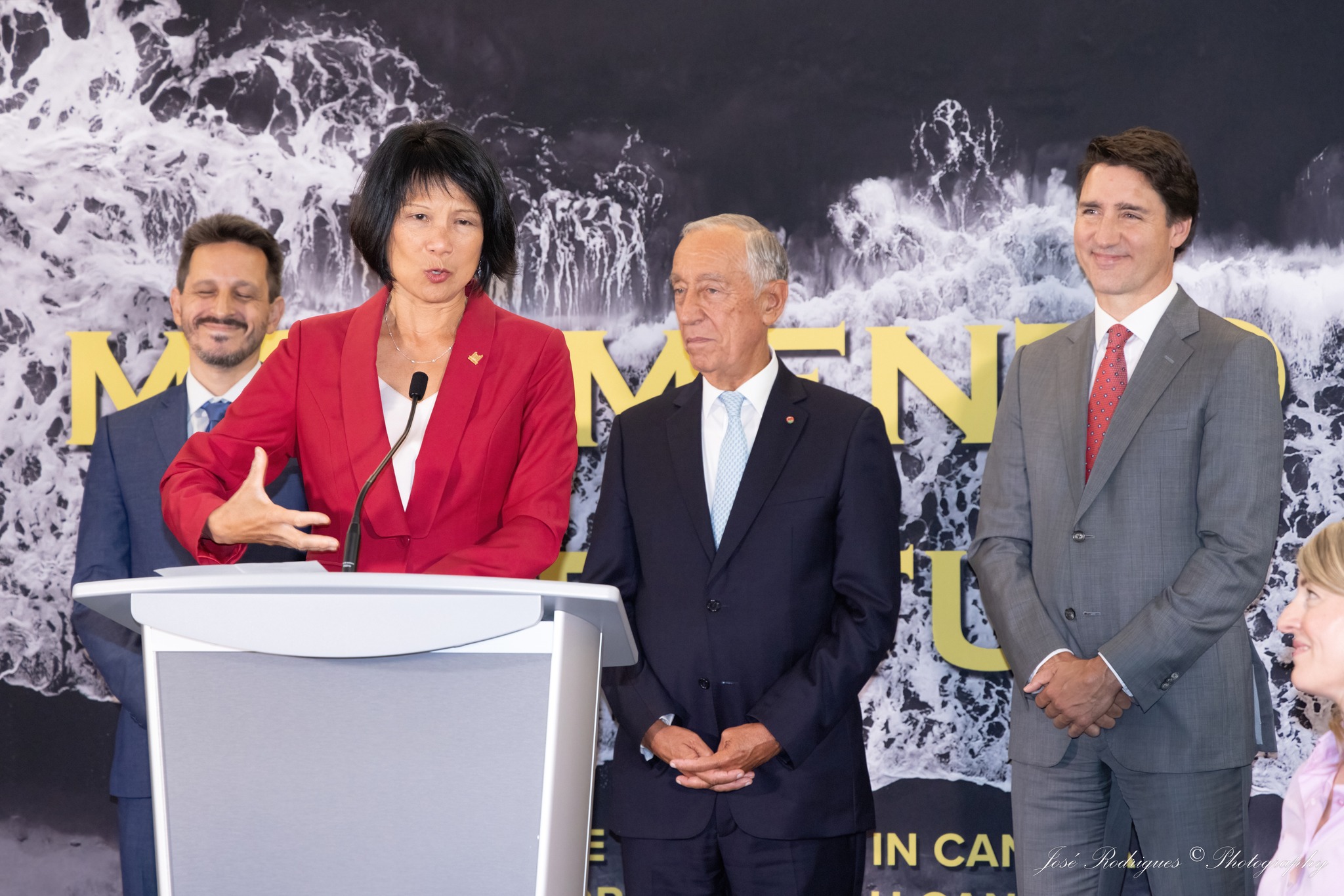 A women speaks at a white podium with three men in suits standing behind her.