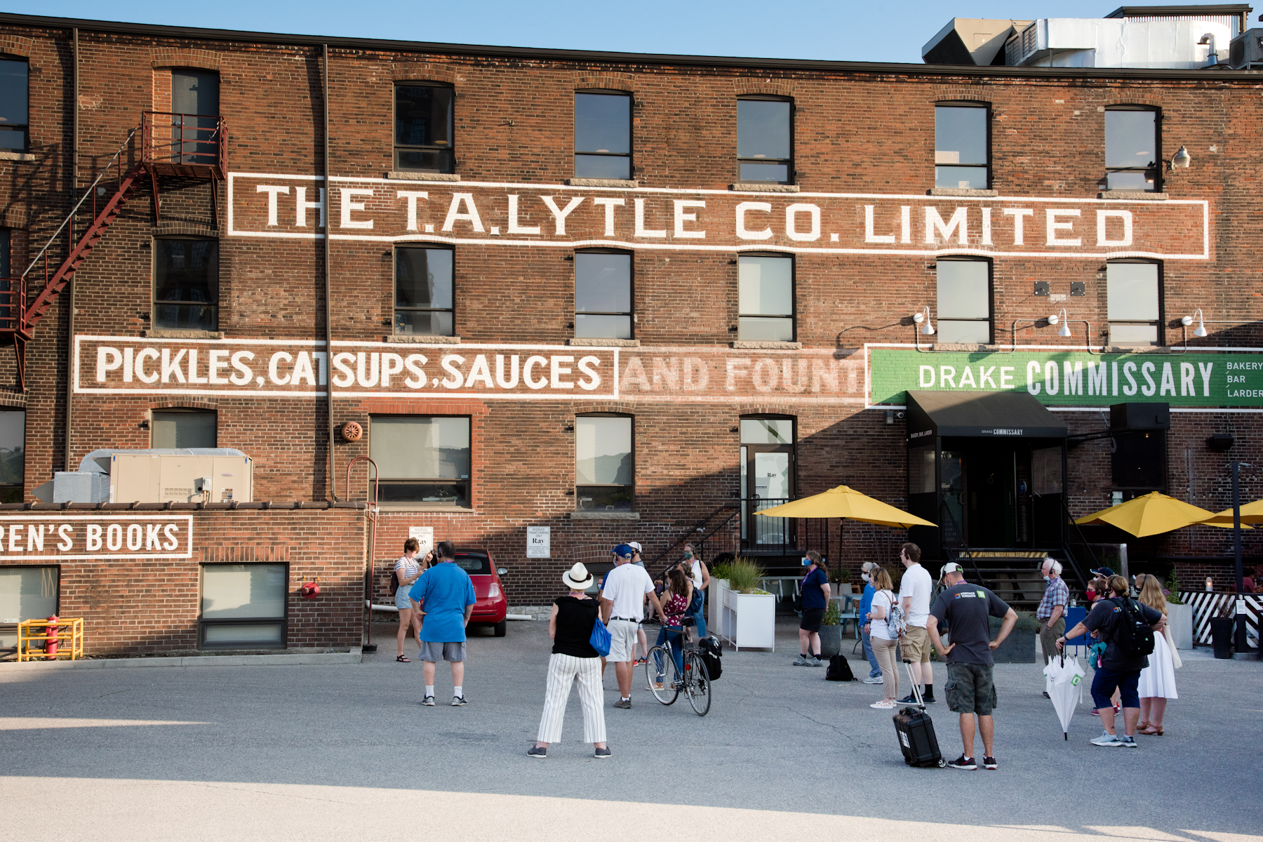 Group of people are gathered outside a red brick building. Text painted in white on side of building reads: "The T. A. Lytle Co. Limited. / Pickles, Catsups, Sauces, and [...] / Fiction, Nonfiction, Poetry, Children's Books".