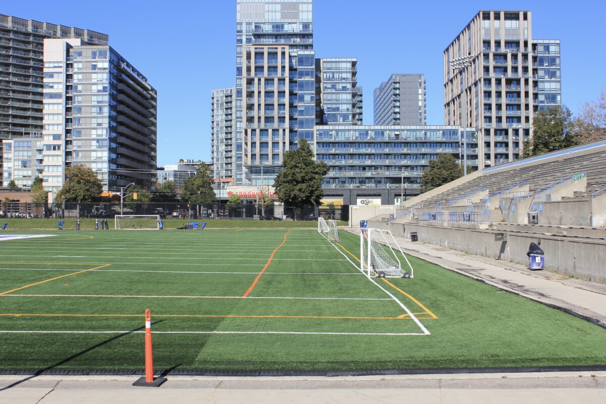 A field and stadium. The seating for the stadium is on the right, while the field (marked with orange and white lines) is to the left. Behind the stadium are a number of tall buildings.