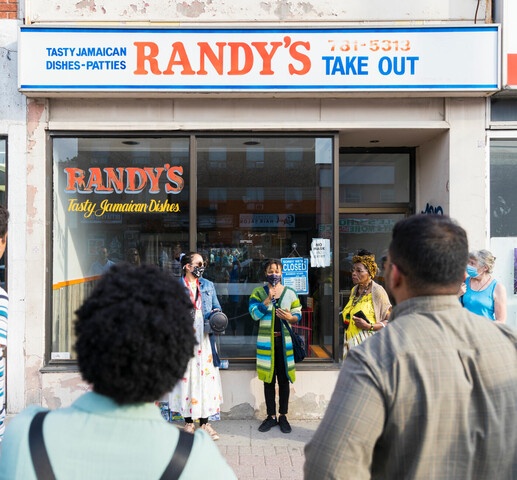 A woman is speaking to a group of people with a microphone and two people are standing beside her in front of a store with glass window panels at the storefront. The store sign above the windows reads " Randy's Take Out" in large blue and red lettering on a white background.