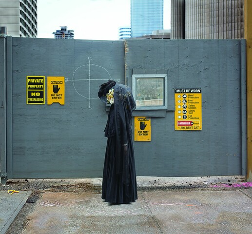 A woman wearing all black, funeral like clothing stands outside a construction site. The wall behind her has a target drawn on in chalk.