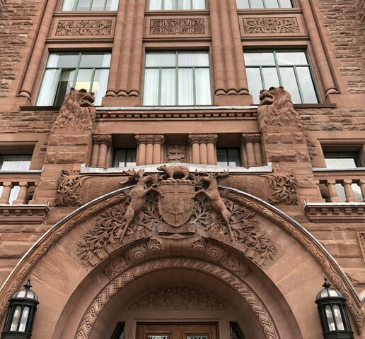 A lower angle image of a red brick structure. There is a stone archway with lions statues on the front of a building. Right below it sits a Coat of Arms with two Caribou facing each other and a bear in between.