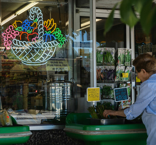 A person standing outside a fruit market, organizing the display in front. There is a neon fruit basket hanging in the shops window,