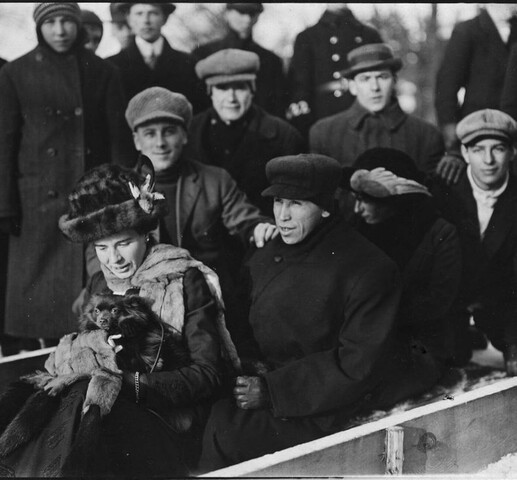 A black and white image of a woman and two men on a toboggan. The woman is holding a small dog.