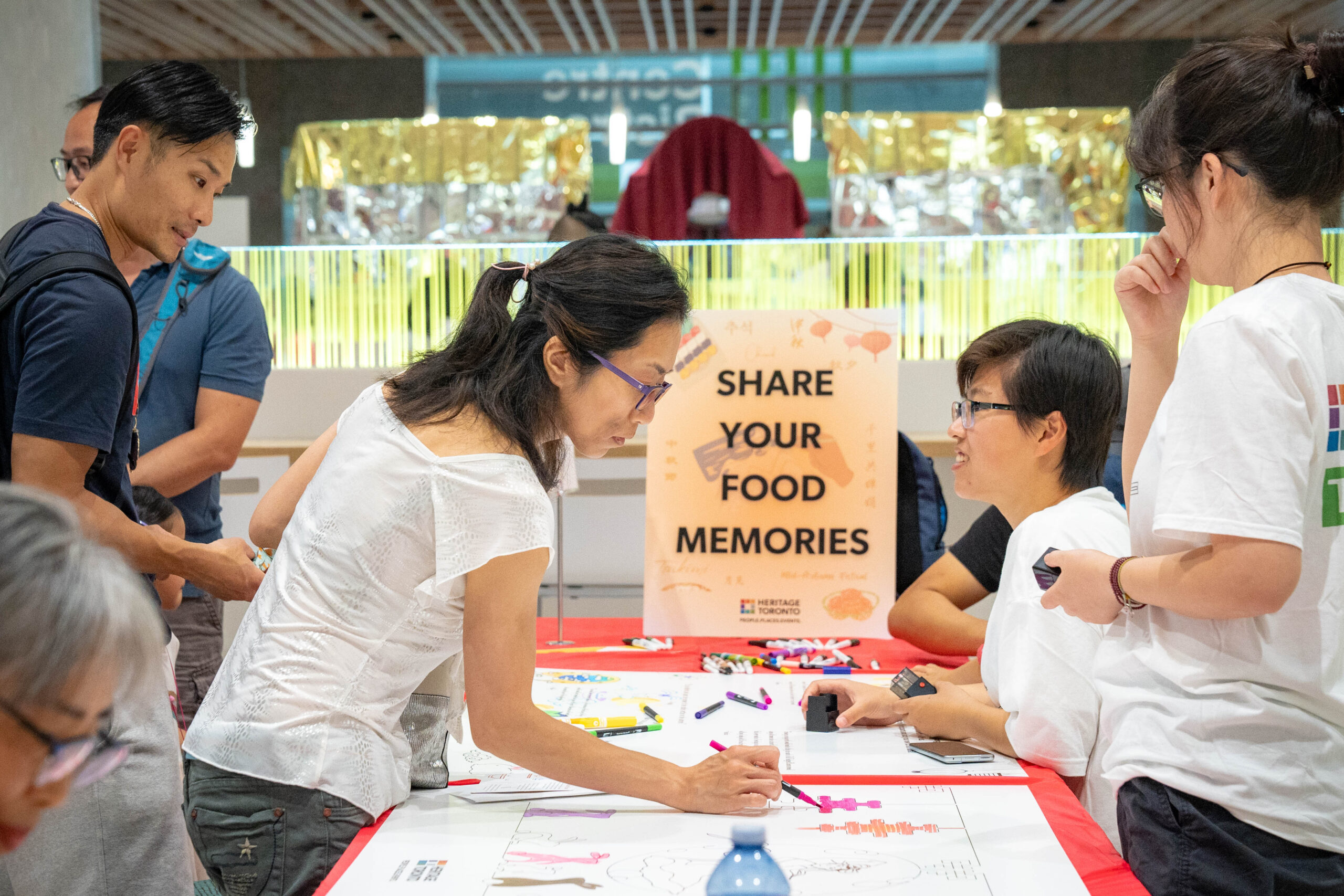 People stand on either side of a long table at the end of which is a sign reading “Share your Food Memories.” One woman is seen writing or drawing on colourful materials that are placed on the table while other people talk and smile.