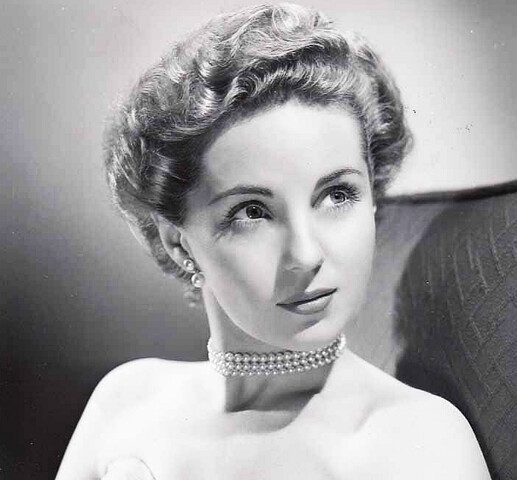 A black and white image of a young woman. She wears earring and a pearl necklace.