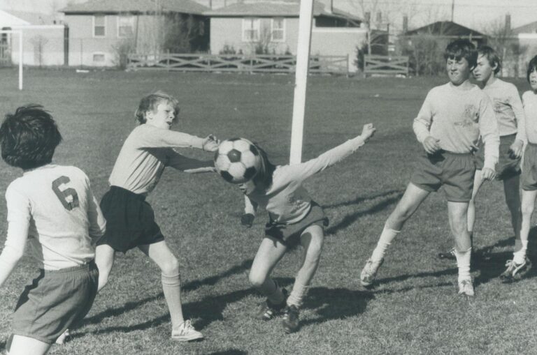 Six children on a field play with a soccer ball. One tries to head the ball.