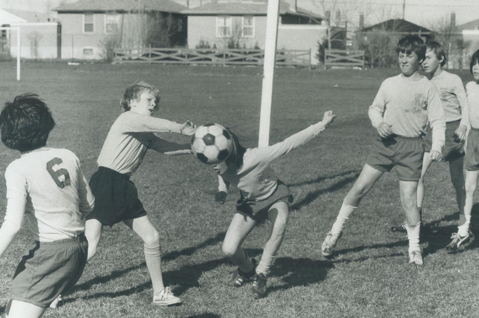 Six children on a field play with a soccer ball. One tries to head the ball.