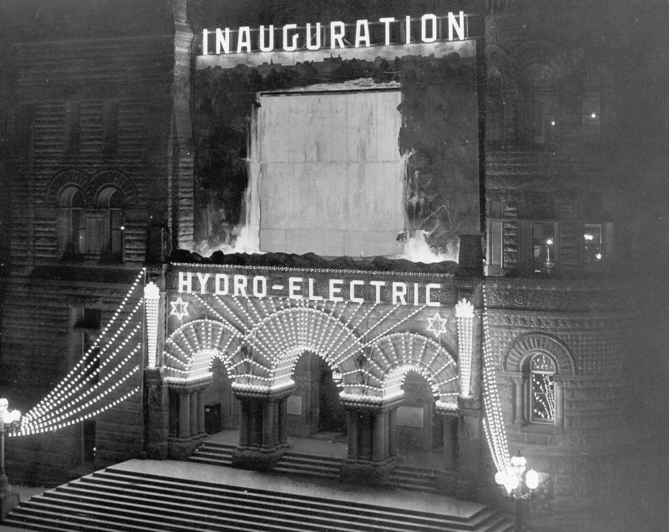 Photograph shows the front of Toronto City Hall at night. The facade is lit up with electric lights reading "Inauguration, Hydro-Electric." Above the main doors of City Hall is a picture of Niagara Falls to show where the hydro-electric power originated.
