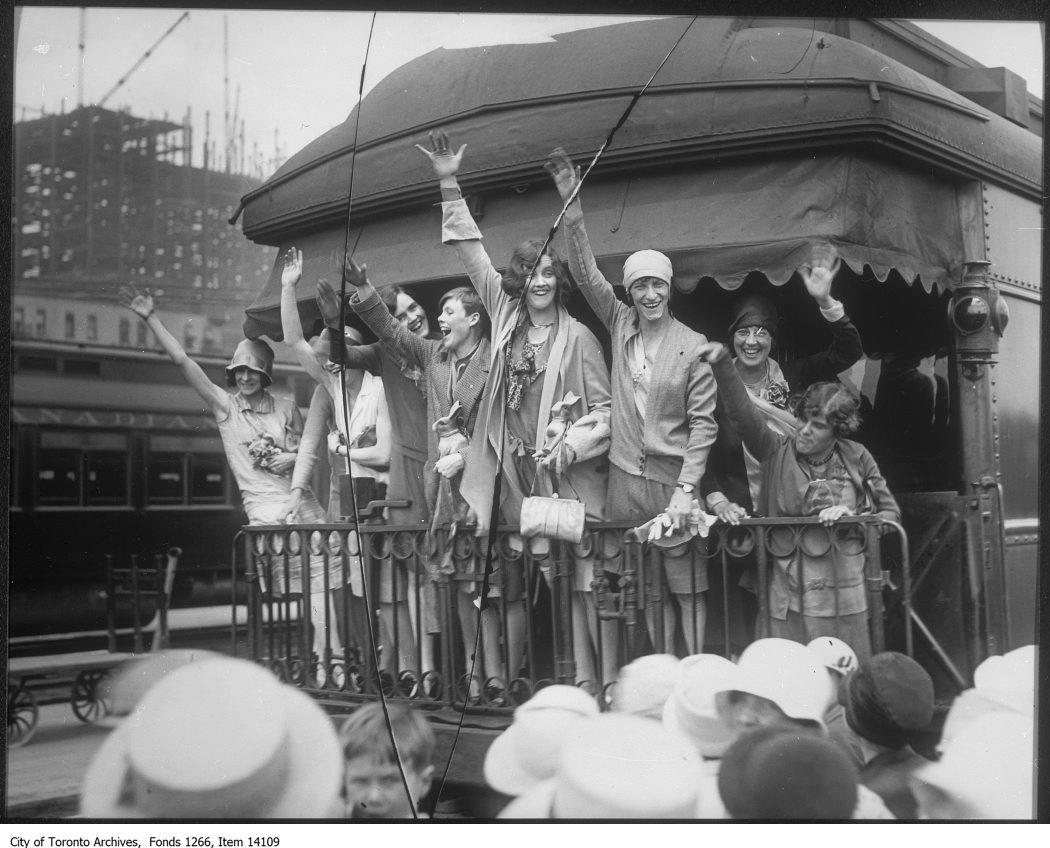 A black and white photograph of eight women standing aboard the caboose of a train in Union Station, Toronto waving to crowds of fans.