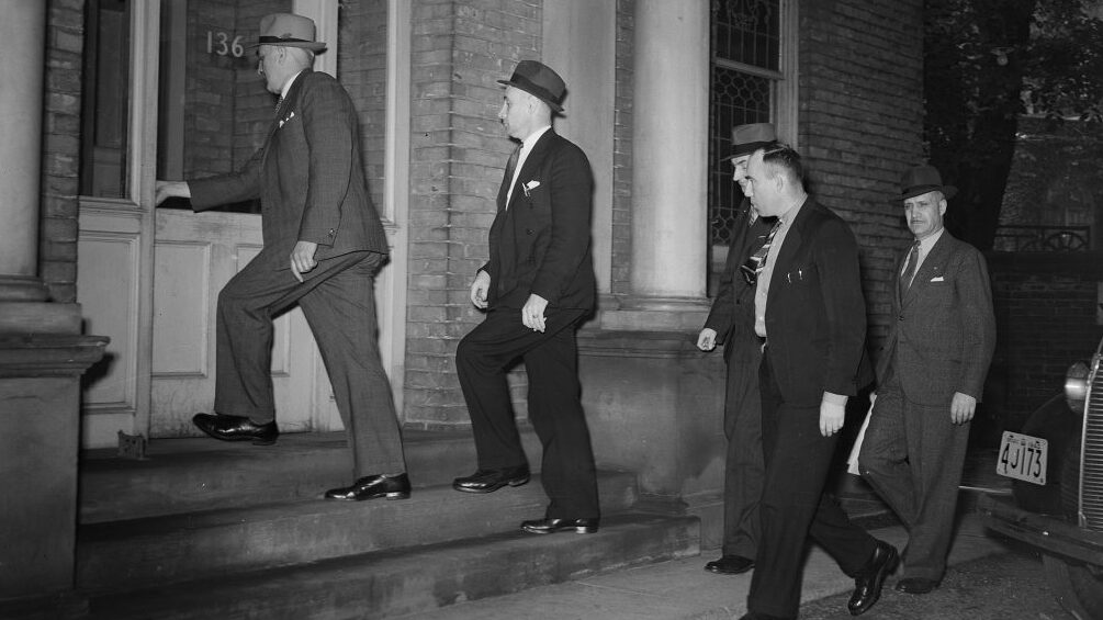 Black and white image of a group of 5 plain clothes police officers walking up to the doors of the Italian Consulate in Toronto.