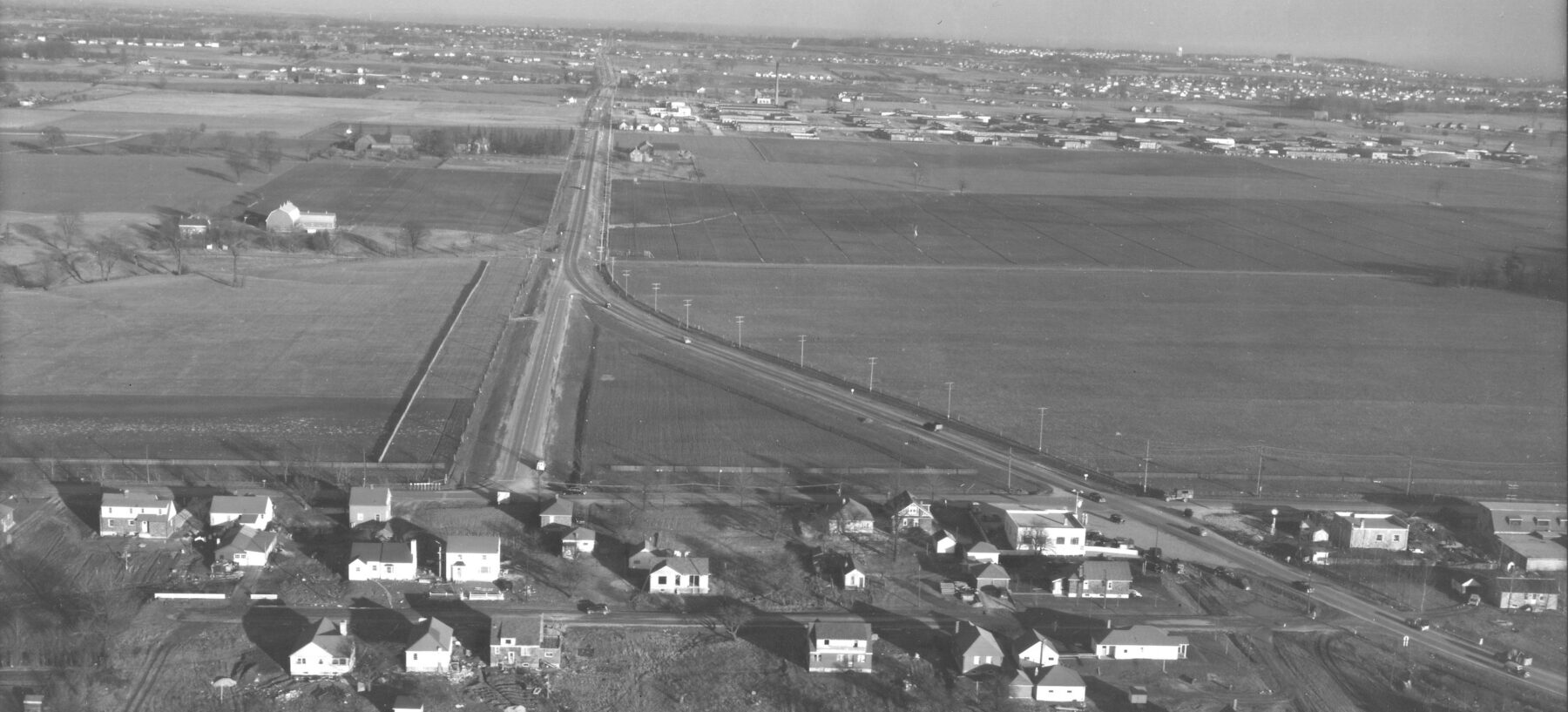 Black and white slightly aerial photograph looking east along Eglinton Avenue East. The area is mostly agricultural with a scattering of houses. A factory is visible in the middle distance.