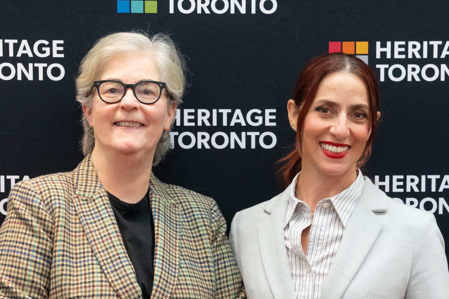 Image of two people - the one on the left wears a beige and black chequered blazer and black top. The one on the right dressed in grey blue blazer and grey blue stripped top. A backdrop behind displays the Heritage Toronto logo.