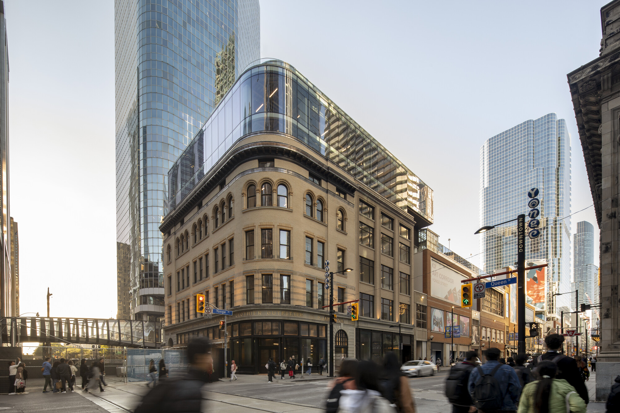 Pedestrians cross a busy Yonge and Queen intersection in front of a restored heritage building topped with a modern glass addition.