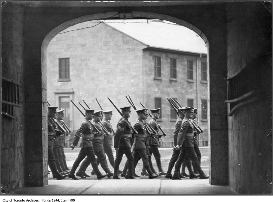 Sixteen male soldiers in uniform with rifles march four astride past an arched gateway. A two-storey stone building is in the background.