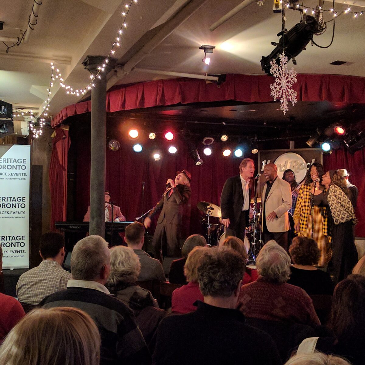 Colour photo taken from the back of the room showing the audience watching a group of musicians performing on stage. A Heritage Toronto banner can be seen on the left hand-side.