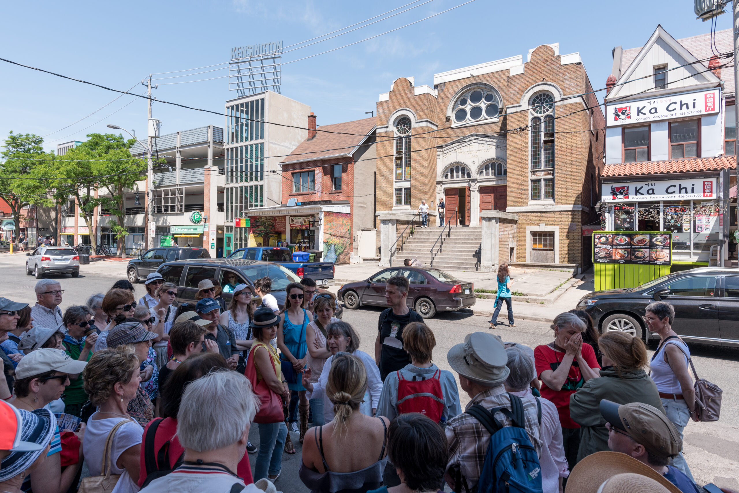 A large crowd stands on the side of the street looking across to a beige brick multi-storey building, which appears to have a high double door entrance accessed by multiple stairs.