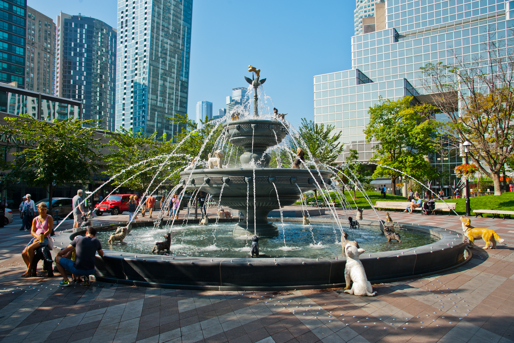 Image of a working fountain surrounded by dog sculptures which spew water out of their mouths.  On the left, two people are sitting on the edge of the fountain. In the background is a bright blue sky framed by several tall buildings and several trees in full green leaf.
