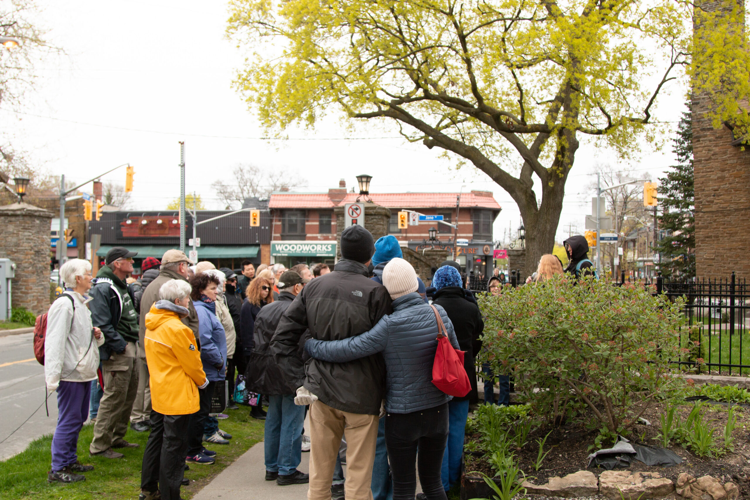 A group of people appear along a sidewalk with a central pair embracing each other. In the background storefronts appear with a stone gate pillar visible on the far left.