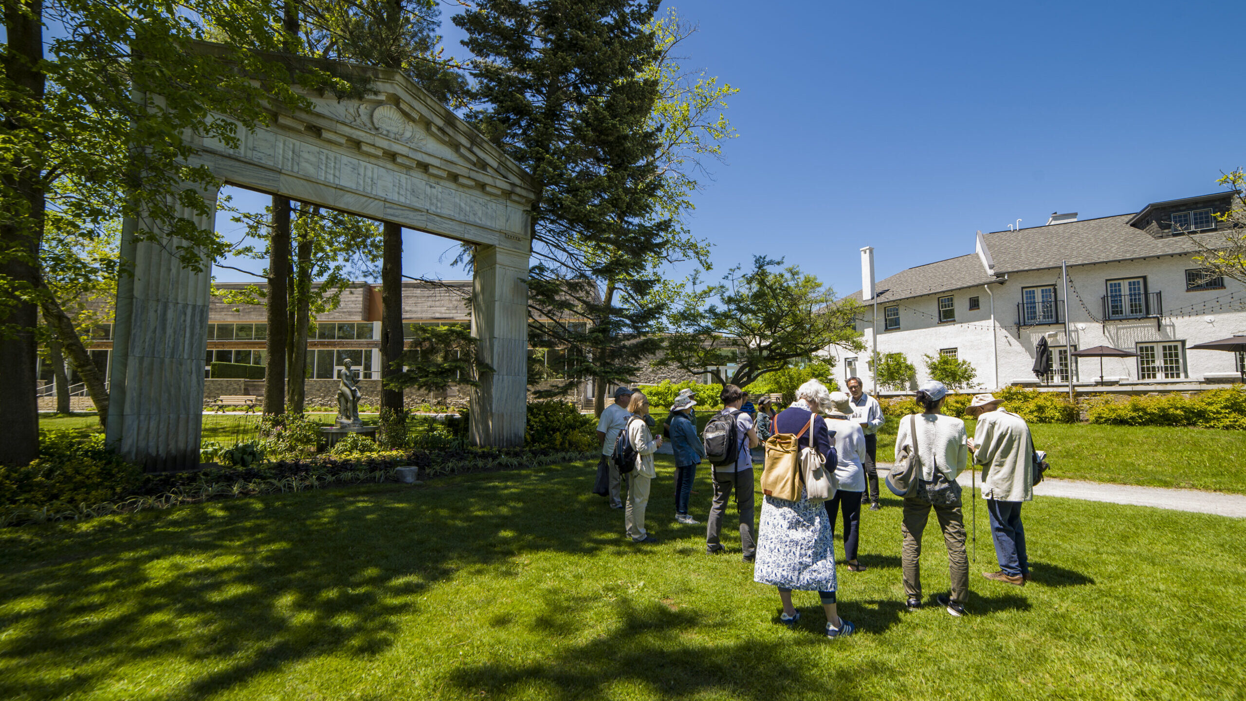 Several people stand in park-like setting. To the left, a large archway appears surrounded by trees. In the background, buildings are visible.