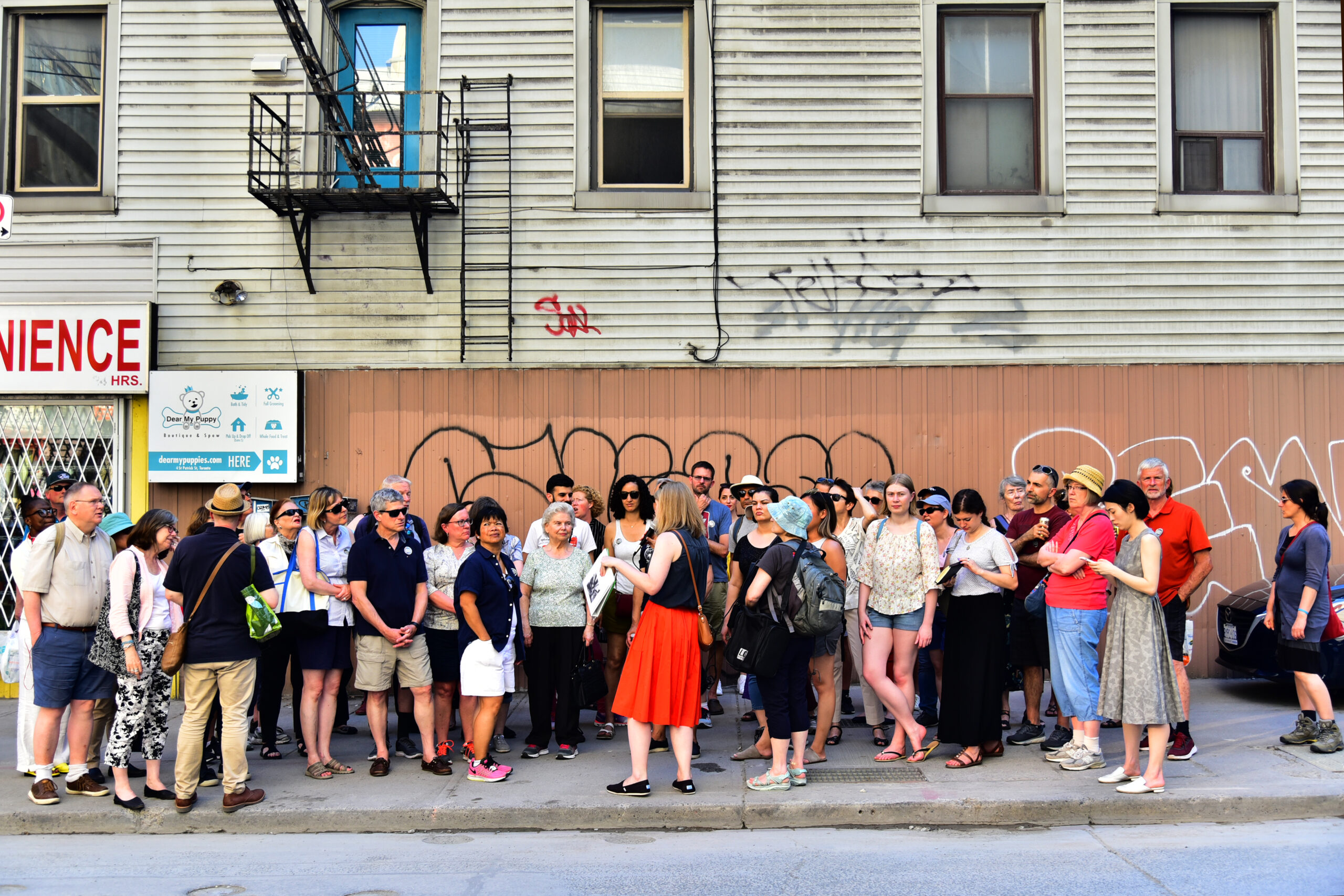 A large group of people stand on a sidewalk. Behind them is a building with some graffiti and storefront signs partially visible, along with a fire escape ladder system.