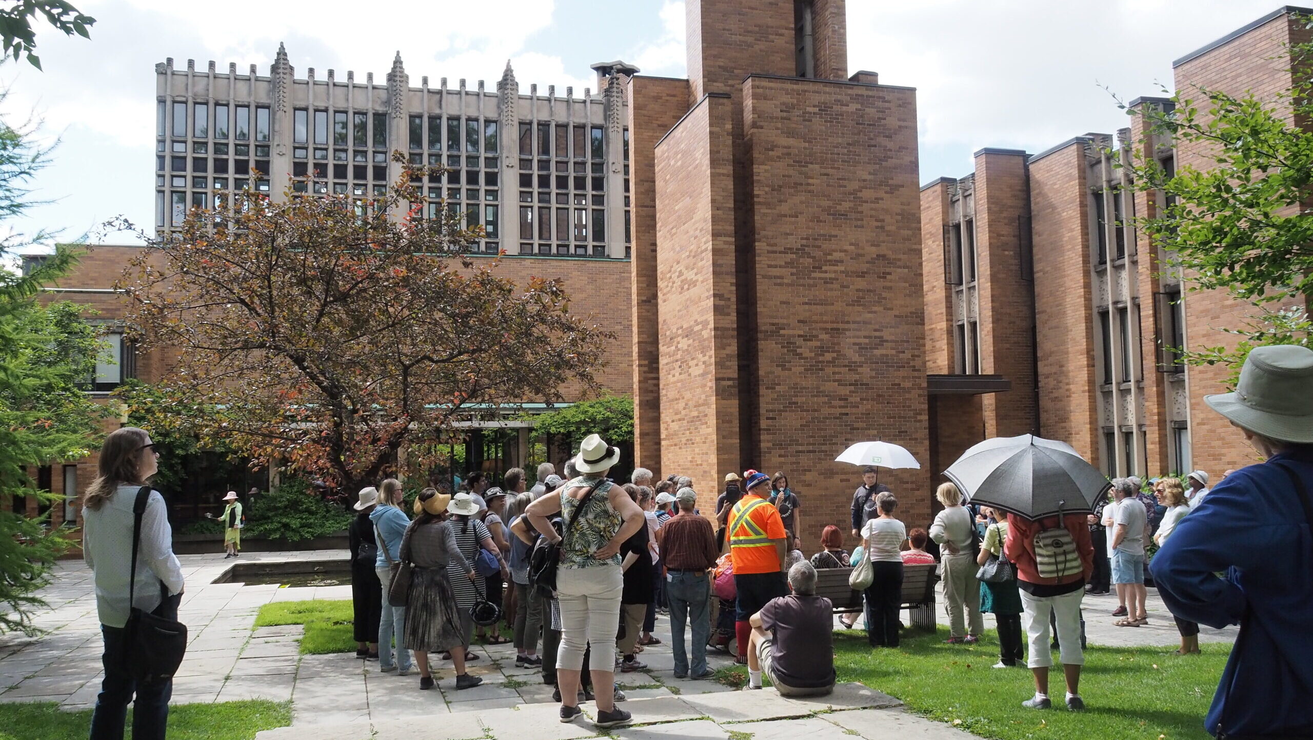 A group of people standing with their backs to the camera are looking at an orange brick building. In the middle of the photograph is a tall square tower flanked by two shorter brick and glass wings.