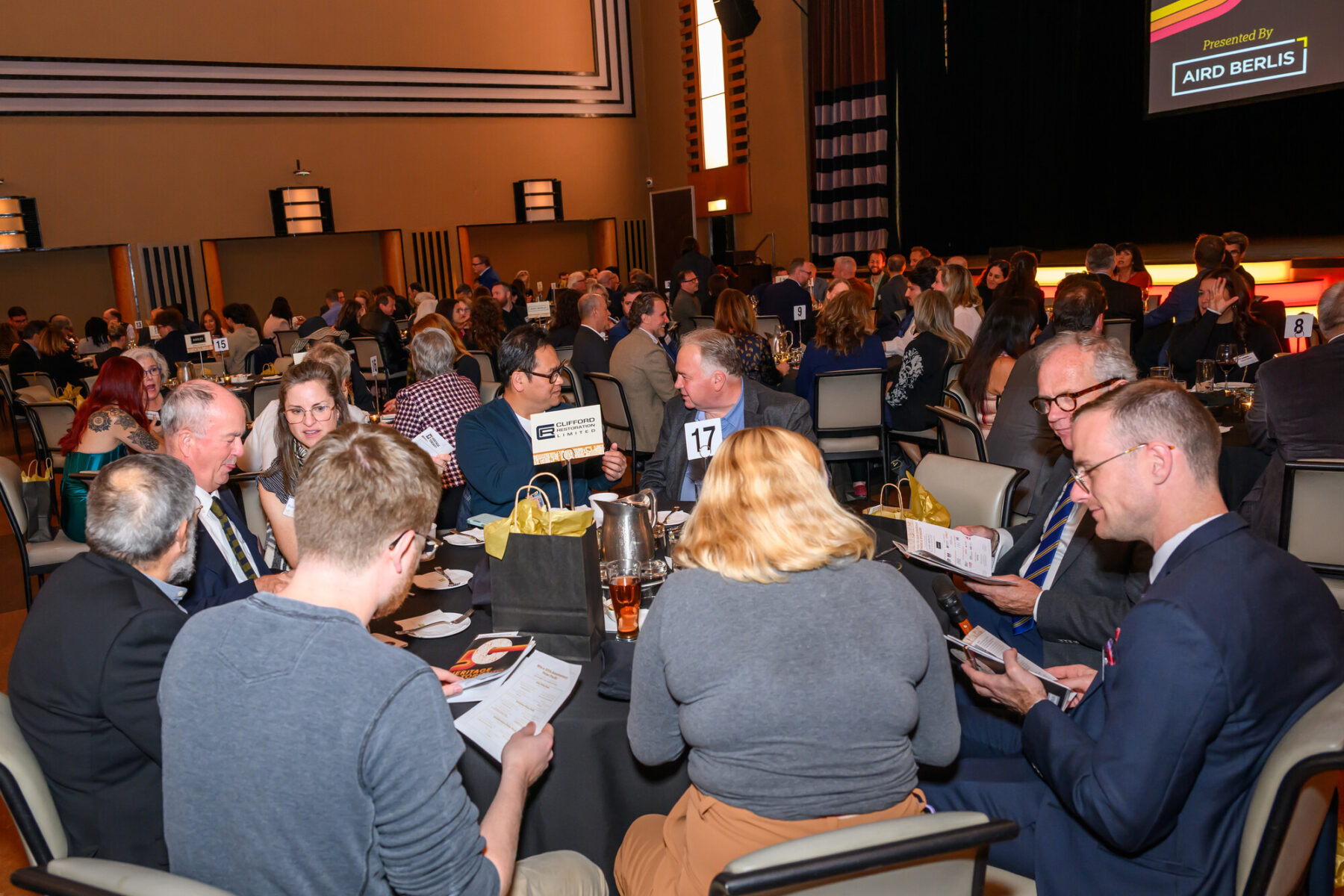 Nine individuals seated at a table. Food and drinks are seen on the table, along with a sign with the number 17 and the logos for the event sponsor Clifford Restoration Limited.