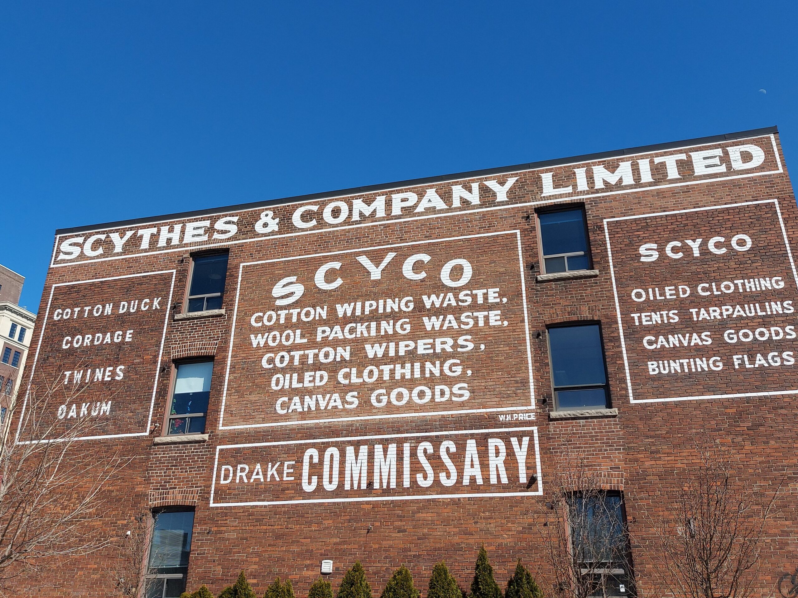 Photo of a red-brick building that has white writing painted on it. The writing reads, "Scythes & Company Limited. Scyco: Cotton wiping waste, wool packing waste, cotton wipers, oiled clothing, canvas goods / Drake Commissary." The sky above the building is bright blue.