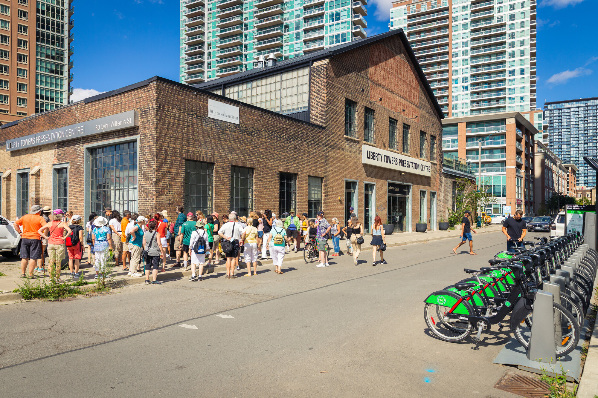 Group of people stand outside a brick warehouse building behind which modern skyscrapers are visible.