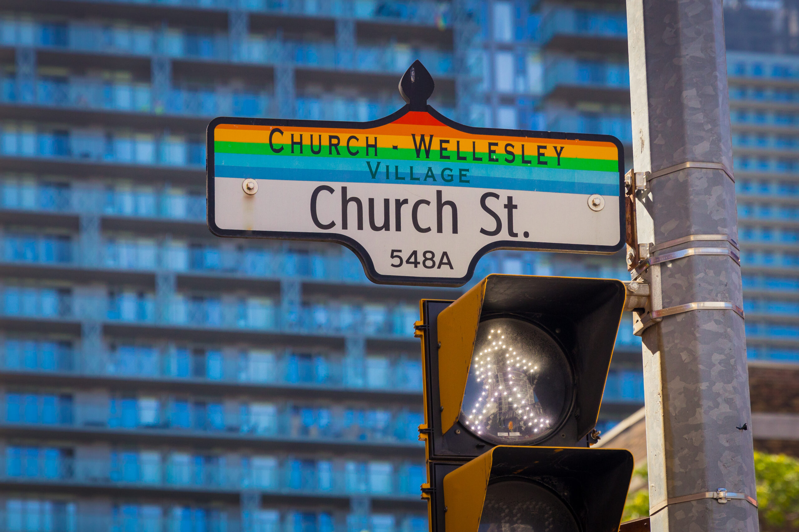 Image of a street sign which reads Church-Wellesley Village / Church St. / 548A. The sign appears above a walk signal with pedestrian figure activated. In the background a multi-storey apartment building appears.