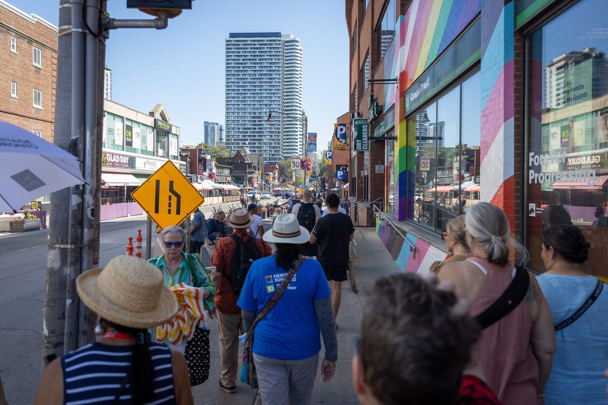 Group of people walk down a street. Brick walls on the right are painted in rainbow colours.