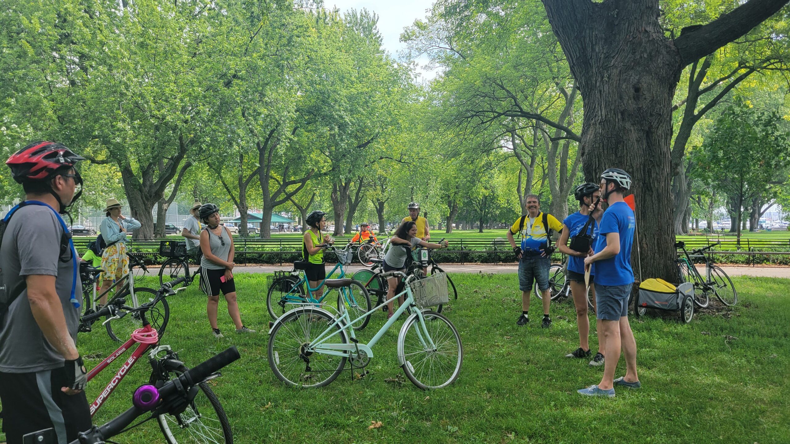 A group of people with bikes stand gathered in a green park around a man with a blue tshirt wearing a bike helmet and holding a clipboard. There are trees in the background and a view of the lake.