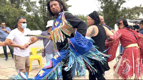 A man in colourful cultural clothing moves in the direction of the camera. His right arm is linked with a woman, and her right arm is linked with another woman. In the background, another line of people with their arms linked faced toward the three people in the center of the image.