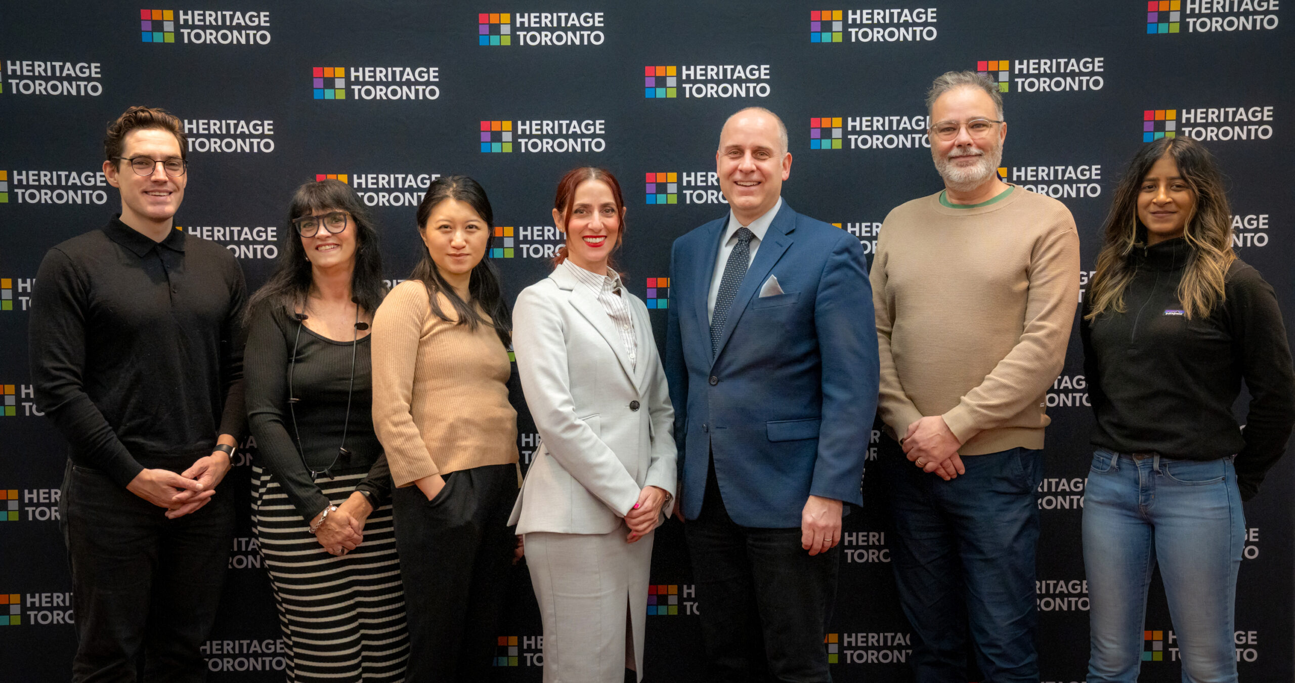 Seven people stand in a row, smile and face the camera. A backdrop behind displays the Heritage Toronto logo.