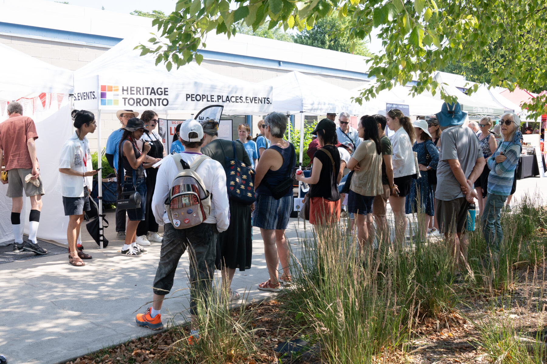 Near a tent with the Heritage Toronto logo, a large group of people gather facing the tent. Some people are situated in front of the tent, addressing the crowd.