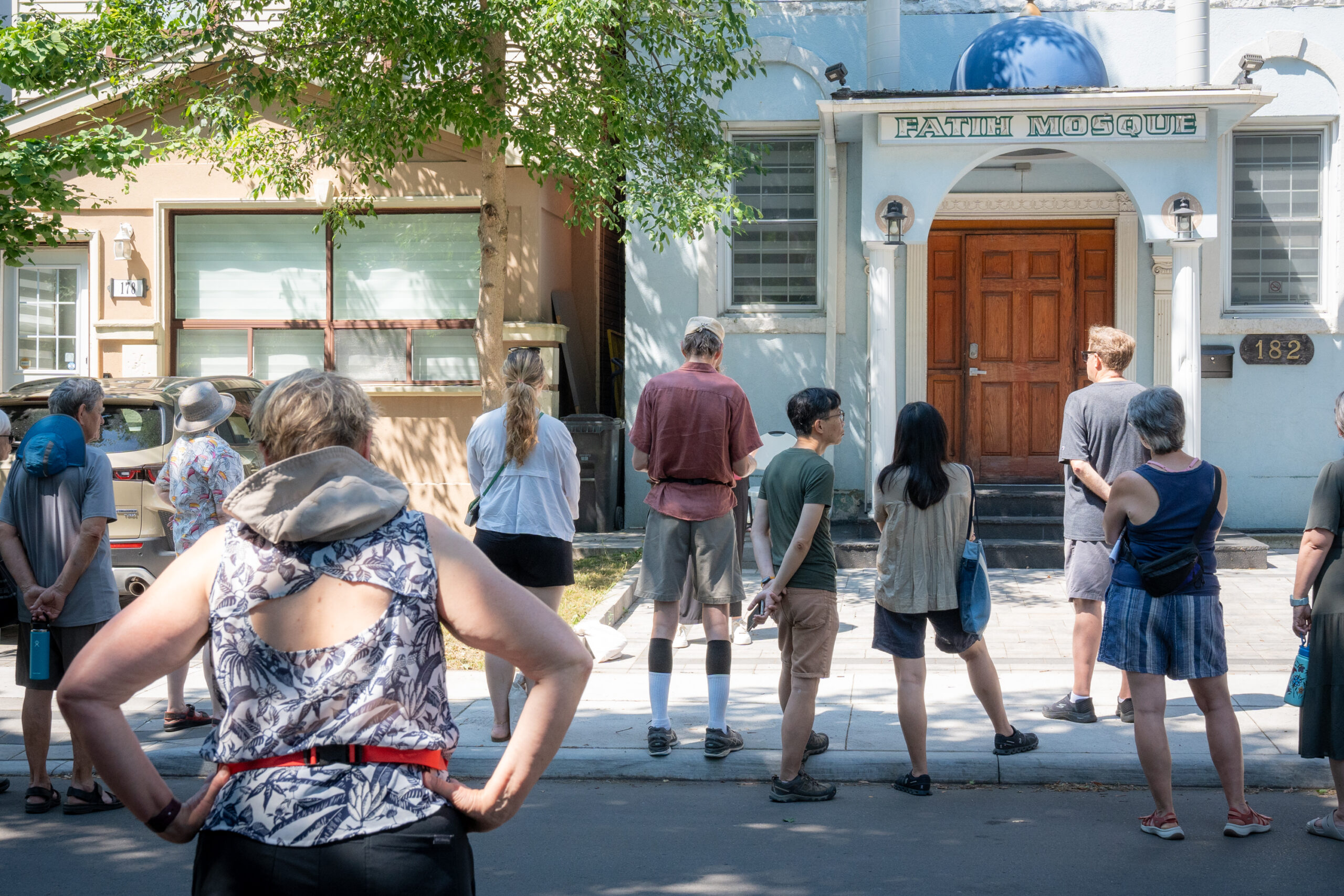 Ten people gather on the pavement in front of a small, white building with a blue, circular architectural structure and a brown, wooden door. The building has a sign that reads "Faith Mosque".