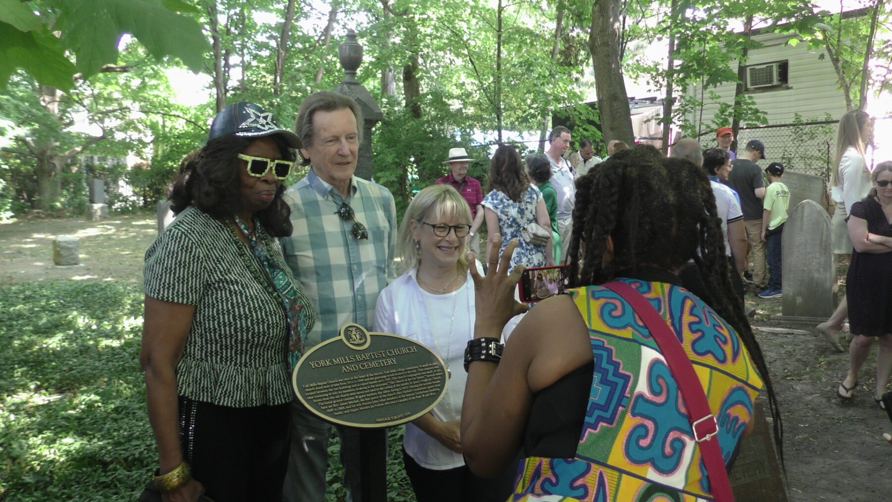 Three people stand in front of a gold and black plaque and pose for a person who is taking a photo of them using a handheld mobile phone. In the background are grass, trees, fences, and a group of people standing around.