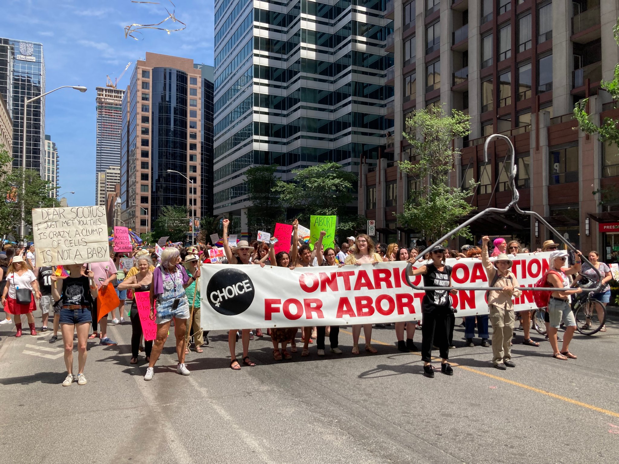 A large group of people, mostly women, walk down a city street. At the front of the group, individuals carry a large white banner with red lettering that reads "Ontario Coalition for Abortion Clinics." In front of the banner, three individuals stand holding an oversized coat hanger. Behind the banner, other individuals can be seen, many of whom carry signs.