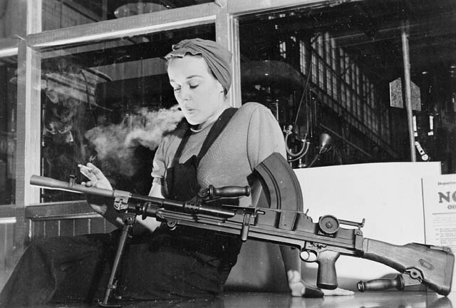 A black-and-white image of a woman in a factory. She is wearing overalls and a scarf on her head. In her right hand is a lit cigarette , which she is smoking. On her lap is a machine gun.