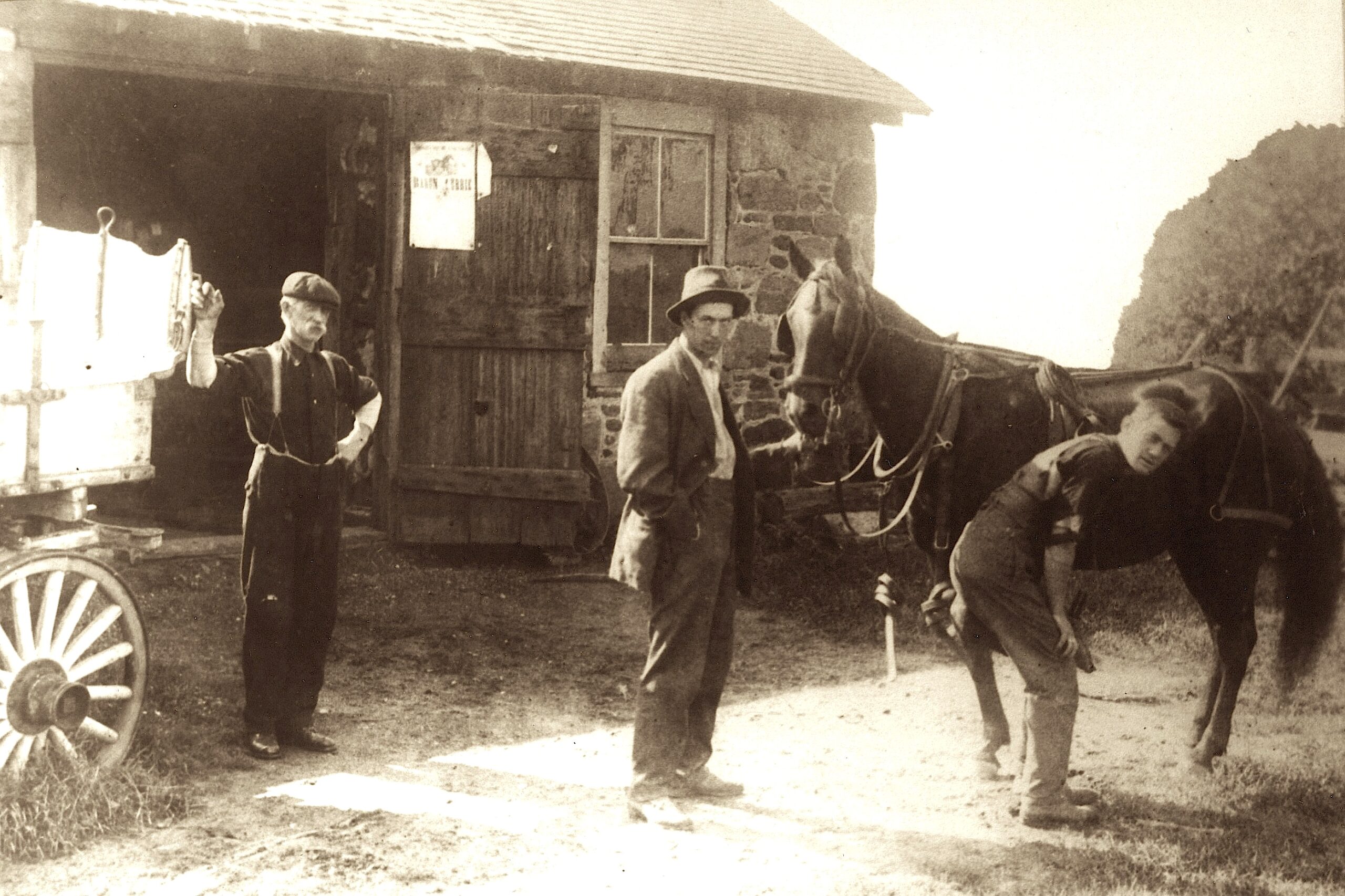 Black and white photo of three men standing outside of a wooden building with large double doors. One man is leaning against a carriage, another holds the reigns of a horse and is facing the camera, and the other man is bent over tending to one of the horse's hooves but is facing the camera.