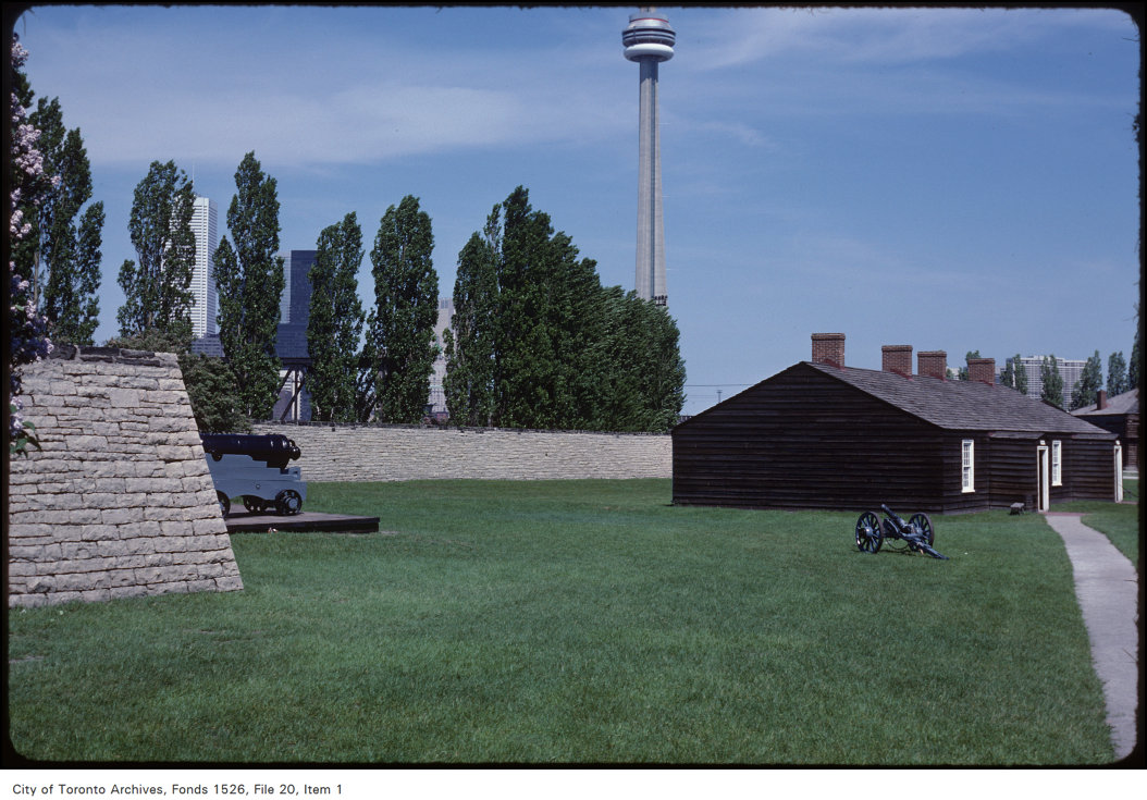 A small wood building sits on a lawn surrounded by a stone wall. Two old cannons are nearby. CN Tower is in the background.