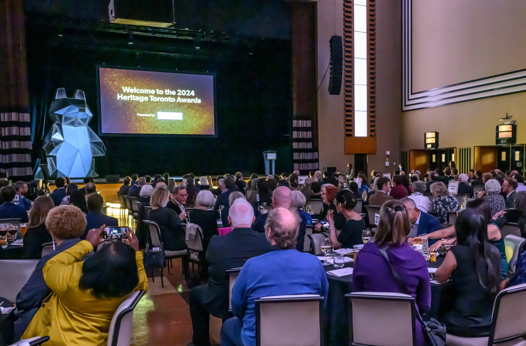 A photo taken looking towards a stage with a podium (to the right), a large animal sculpture (to the left) and a central screen projection with the words "Welcome to the 2024 Heritage Toronto Awards" visible. In the foreground, groups of people sit at round tables.
