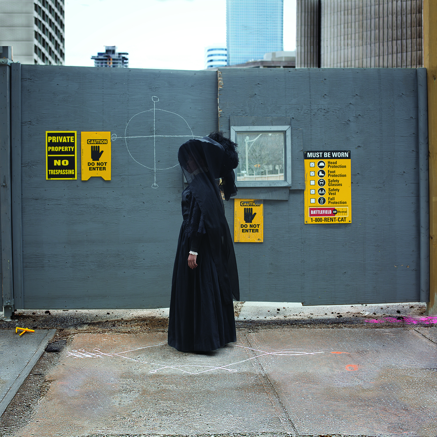 A woman wearing all black, funeral like clothing stands outside a construction site. The wall behind her has a target drawn on in chalk.