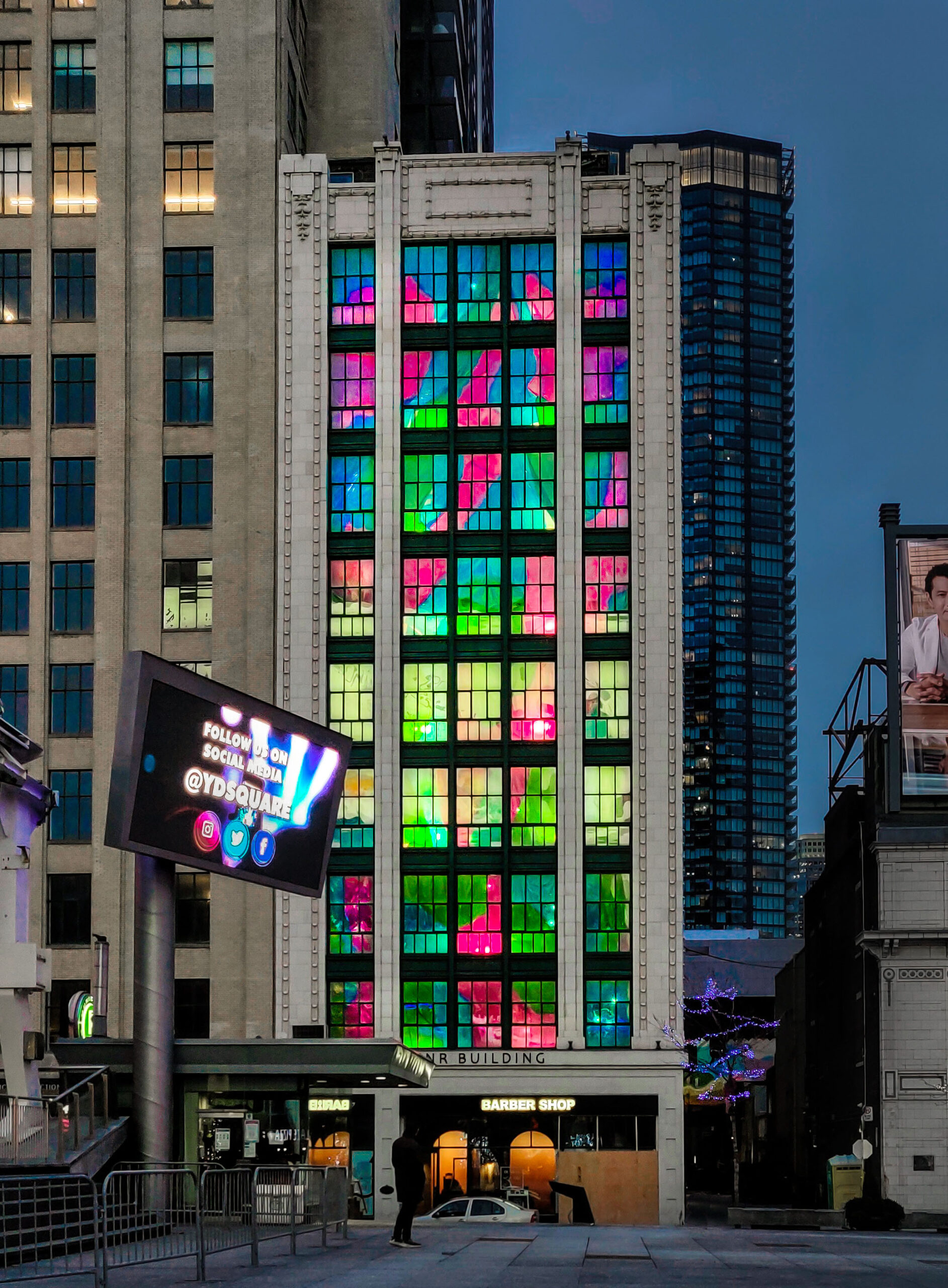 A nine-story light grey building with bright, colourful windows and an underground parking entrance. A bright billboard is in front of the building to the left.