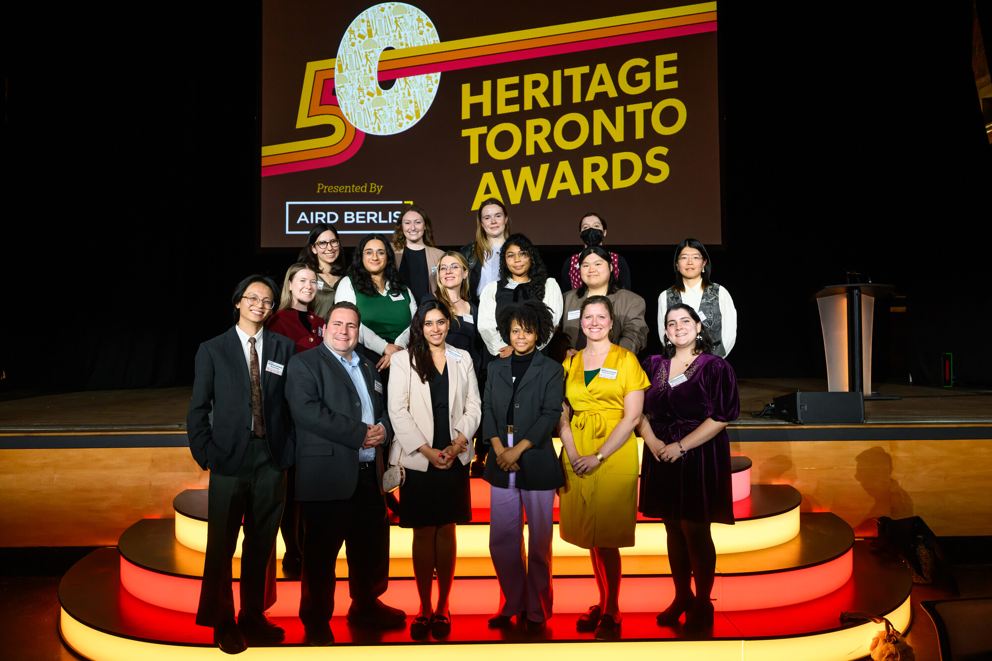 Image of 16 people. They stand in three rows on steps to the stage. They are dressed in business casual wear. On stage a screen displays a slide with the Aird & Berlis logo and text reading: 50 Heritage Toronto Awards.