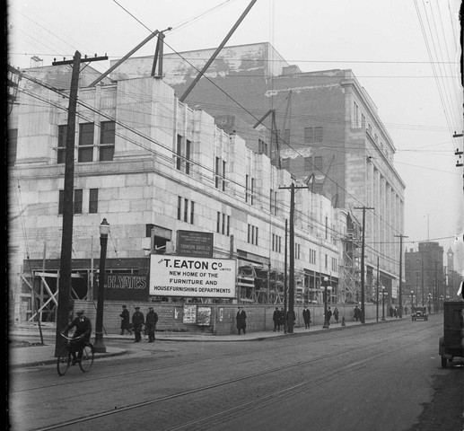Black and white image of Eaton building. One section is four storeys high, and the other is seven storeys high. On the corner is a sign for T. Eaton Co, which says Eaton's is the new home for furniture and home furnishing departments.