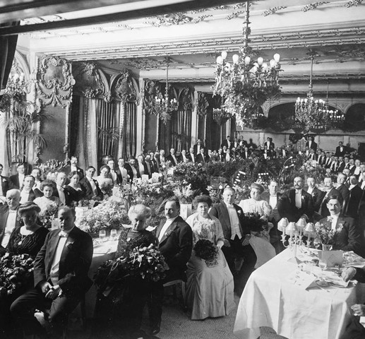 Black and white image of a crowded ballroom with many patrons who are all formally dressed seated for a meal.
