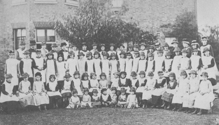 30-40 young girls of different ages are wearing white aprons and hats seated for a group photo outdoors.