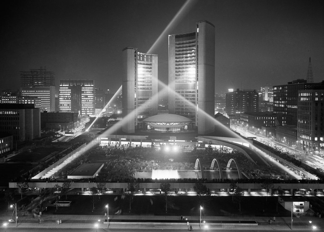 Wide view of a Neo-Expressionist Modern building illuminated by searchlights. It is facing a crowded public square.